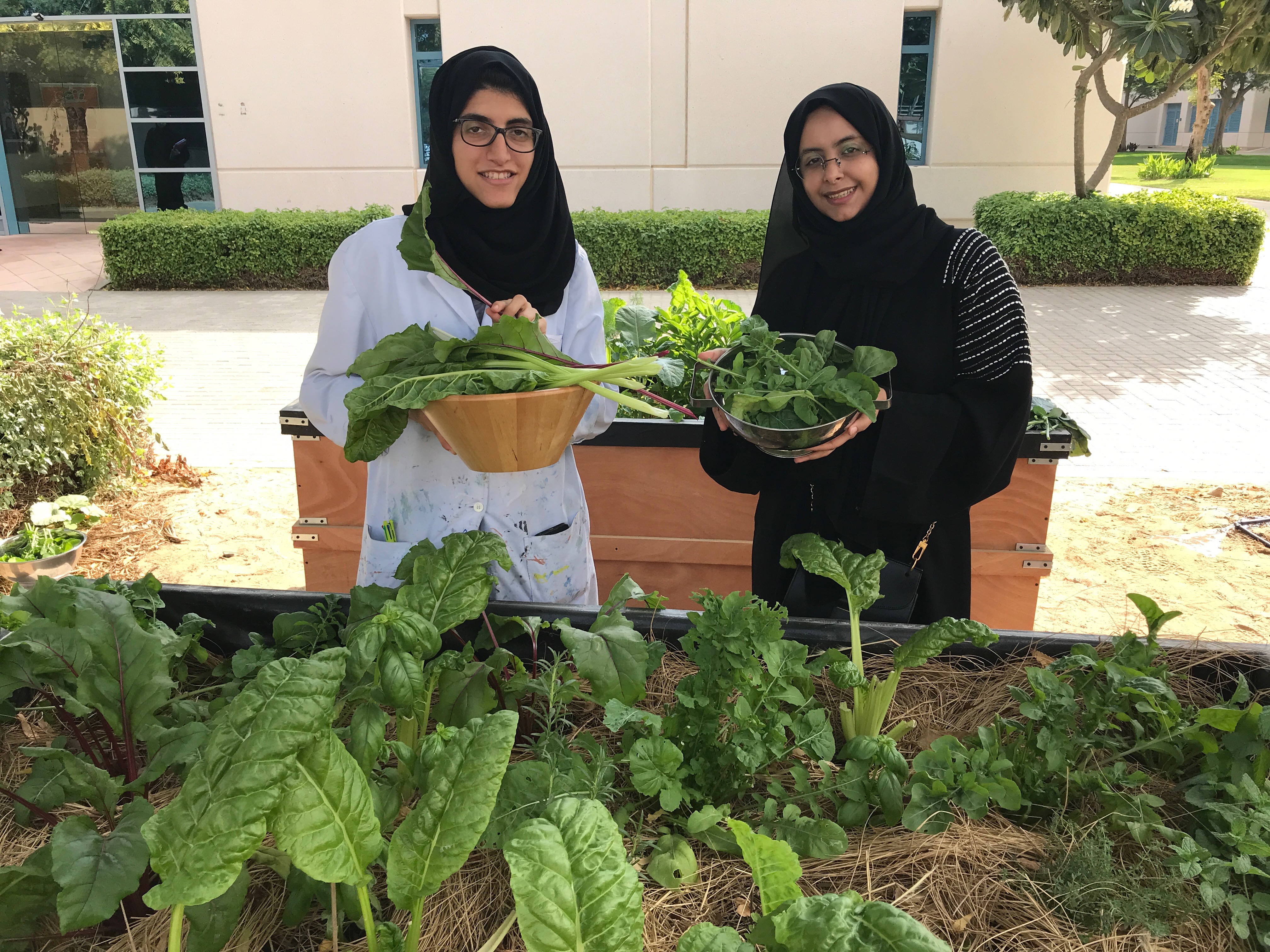 Two students holding bowls of freshly harvested greens beside the raised beds of the KULNA garden