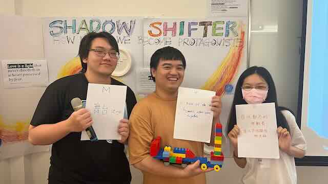 Three young people holding hand-written cards and a LEGO model in front of the hand-drawn Shadow Shifter wall