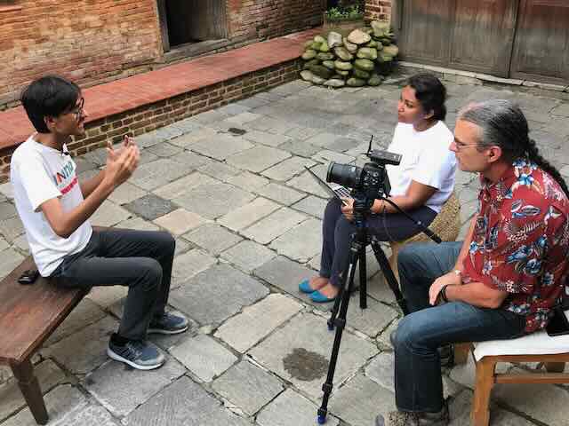 A young Nepali man being interviewed in a brick courtyard — the film crew seated behind the camera